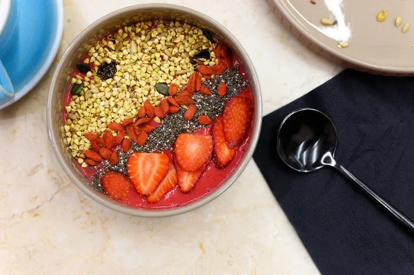 smoothie bowl with blend fruits, strawberry, goji, chia seeds, and green buckwheat granola, and metal black spoon, on the table