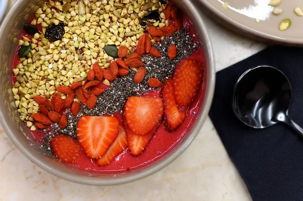 smoothie bowl with blend fruits, strawberry, goji, chia seeds, and green buckwheat granola, and metal black spoon, on the table