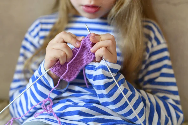 kid girl knitting with needles in home interior on background of gray concrete wall. the concept of teaching and studies at home during the quarantine