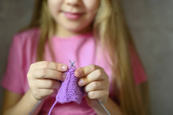 kid girl knitting with needles in home interior on background of gray concrete wall. the concept of teaching and studies at home during the quarantine