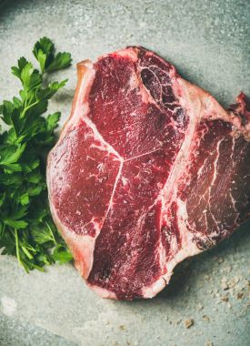 Düz-lay ham pişmemiş kuru-yaşlı t-bone sığır eti biftek taze yeşil maydanoz ile Başbakan ve tuz metal arka plan üzerinde üstten görünüm füme. Sağlıklı protein et yemeği kavramı