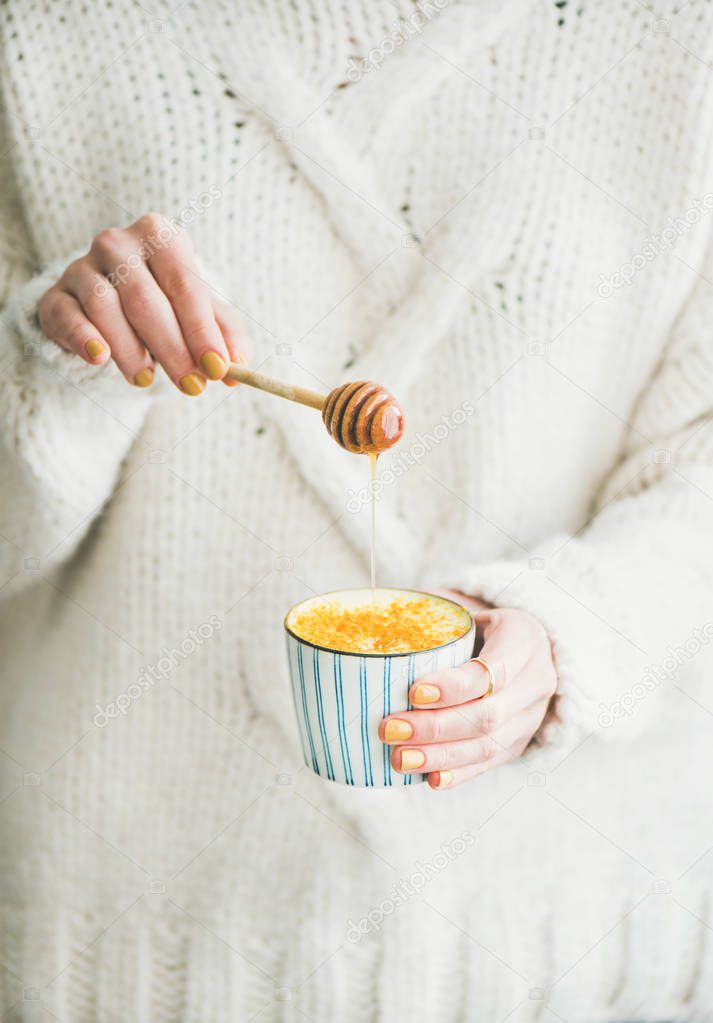 Healthy vegan Ayutvedic hot drink. Female in white loose sweater holding cup of turmeric latte or golden diary-free milk with honey in hands