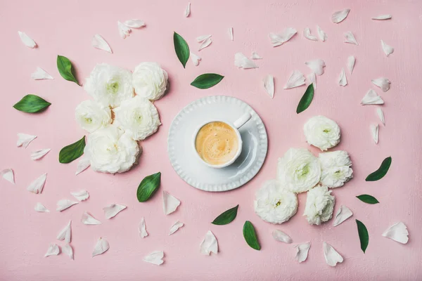 Spring morning concept. Cup of coffee surrounded with white ranunculus flowers and petals over light pink pastel background