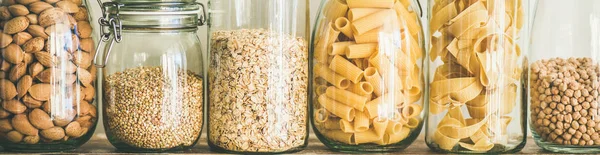 Various uncooked cereals, grains, beans and pasta for healthy cooking in glass jars on wooden table, white background