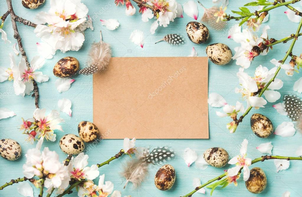 Easter holiday background. Flat-lay of tender Spring almond blossom flowers on branches, feathers, quail eggs and paper in center over blue background