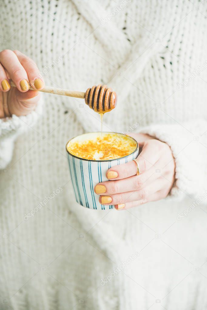 Healthy vegan turmeric latte or golden milk with honey in hands of woman wearing white woolen sweater
