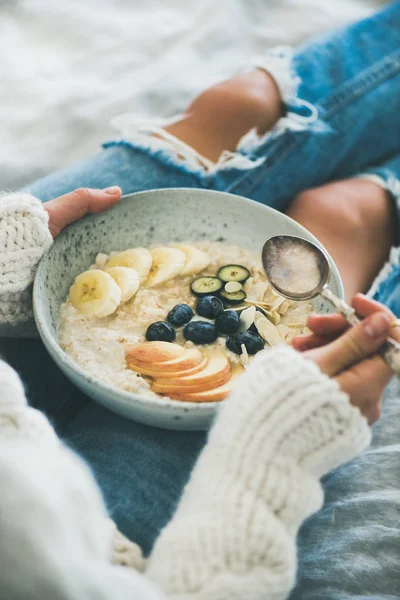 stock image Healthy winter breakfast in bed. Woman in woolen sweater and shabby jeans eating vegan almond milk oatmeal porridge with berries