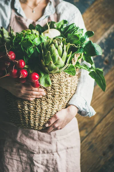 Female farmer wearing pastel linen apron and shirt holding basket with fresh seasonal vegetables in her hands