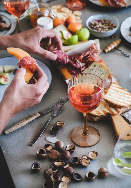 Mid-summer picnic with wine and snacks. Charcuterie and cheese board, rose wine, nuts, olives and fruits and mans hands with food over concrete table background, top view. Family holiday gathering