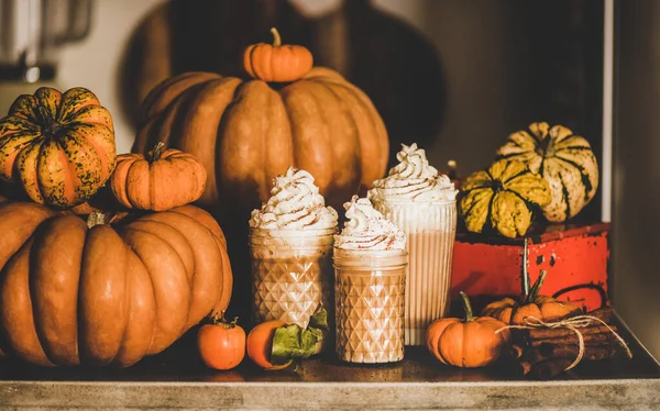 Pumpkin spice latte coffee drink topped with whipped cream in tall glasses among fresh pumpkins, persimmons and cinnamon sticks on table, grey wall background. Seasonal Autumn hot warming sweet drink