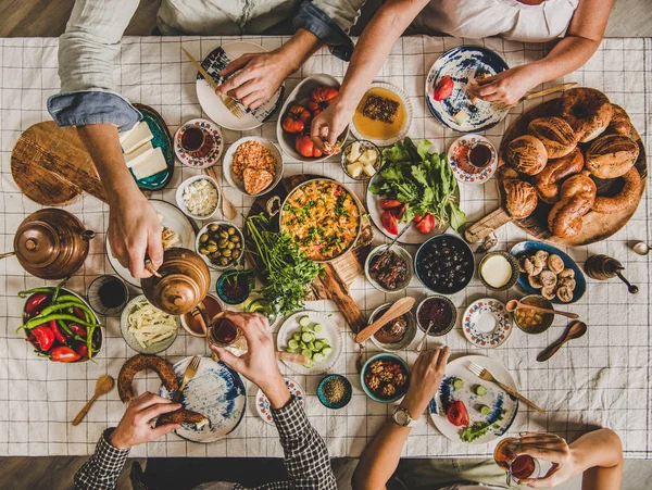 Flat-lay of people sitting at table with Turkish breakfast with pastries, vegetables, greens, cheeses, eggs, jams and tea in tulip glasses and copper teapots over chekered linen tablecloth, top view