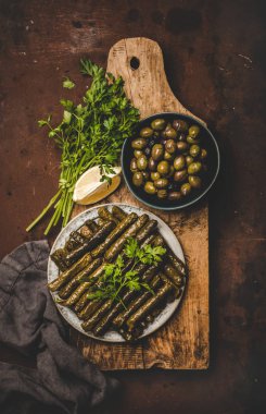 Flat-lay of Turkish traditional grape leaves Sarma or Dolma stuffed with rice and spices garnished with olive oil, olives, lemon and fresh parsley on wooden board over scorched background, top view