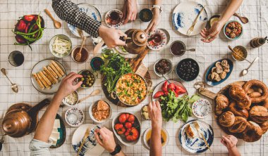 Flat-lay of family having Turkish breakfast with pastries, vegetables, greens, spreads, cheeses, fried eggs, jams and tea in tulip glasses and copper teapots over pastel chekered linen tablecloth