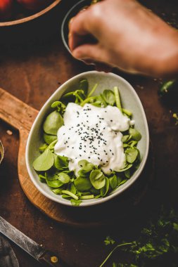 Turkish starter, meze dish. Human hand adding black sesame to bowl of fresh green purslane salad with yogurt over rusty table with vegetables and spices, top view. Turkish, Middle East cuisine
