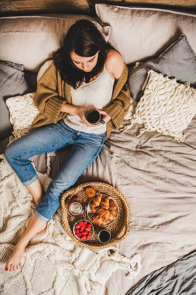 パステルカラーの毛布の上に座っているカジュアルな服の若いブルネットの女性は裸足で 新鮮なクロワッサン イチゴと紅茶 トップビューでベッドで朝食を持っています 快適な生活コンセプト — ストック写真