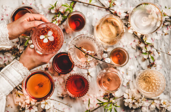 Womans hands over dofferent shades of rose wine in glasses and spring blossom flowers over white background, top view. Wine shop, bar, wine tasting, wine list concept