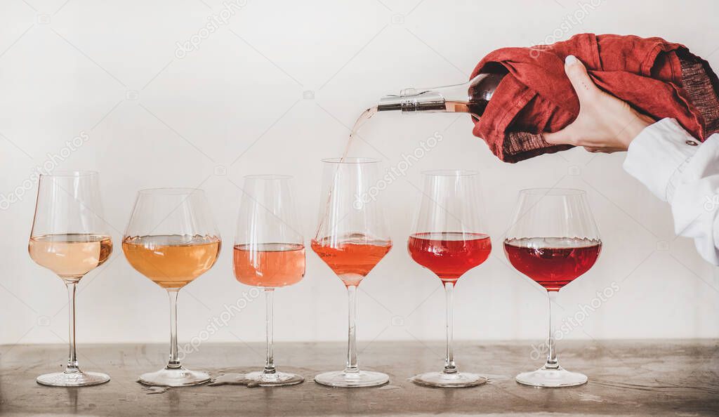 Shades of Rose wine in stemmed glasses placed in line and womans hand pouring wine from bottle to glass, white wall background behind. Wine bar, wine shop, tasting concept