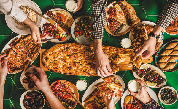 Muslim Ramadan iftar family dinner. Flat-lay of peoples hands over table with Middle East food. Dates, kebab, flatbread, pide, borek, sweet, salad over table, top view. Ramazan fasting Turkish cuisine