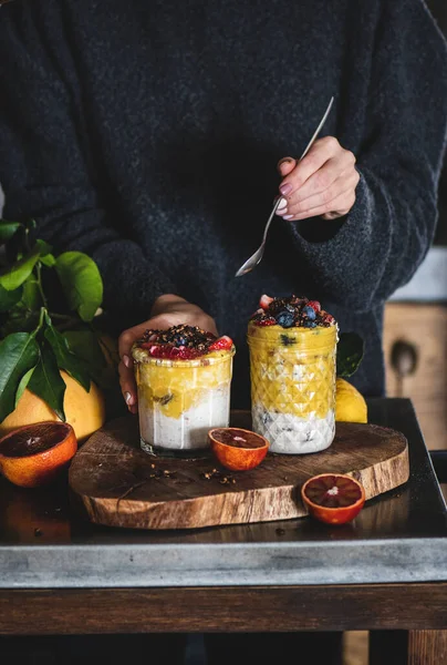 Gri beton mutfak tezgahının üzerinde gece boyu yulaf ezmesi, mango smoothie, karabuğday gevreği ve taze böğürtlen yiyen bir kadın. Sağlıklı, temiz yeme konsepti