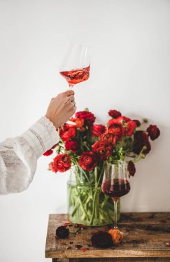 Beyaz duvar arka planında, gül şarabı ve kırmızı bahar çiçeği çiçekli kırsal ahşap masa üzerindeki kadın eli, seçici bir odak noktası. Şarap dükkanı, bar, şarap tatma, şarap listesi konsepti