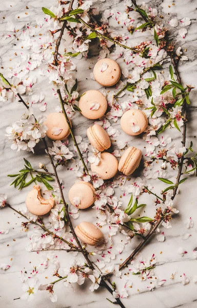 Food texture, wallpaper and background. Flat-lay of sweet macaron cookies and white spring blossom flowers and branches over white marble background, top view, vertical composition