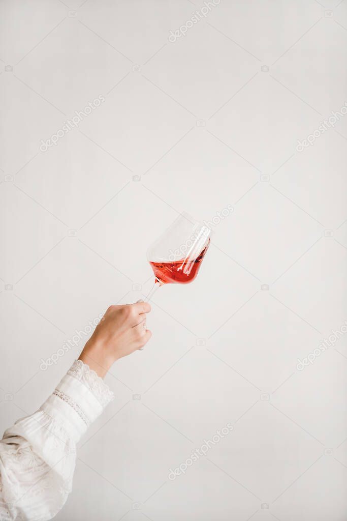 Womans hand in white shirt holding and turning glass of rose wine during tasting over white wall background. Wine shop, wine tasting, bar, wine list concept