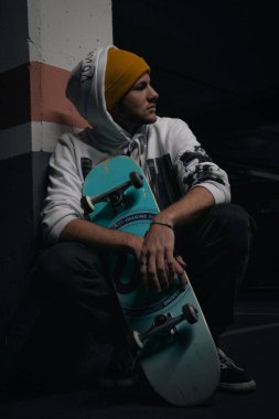 Kaykaylı genç bir adam garajda sweatshirt ve şapka giyiyor.