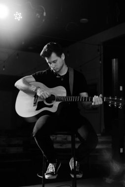 Avrupa görünümlü genç yakışıklı bir adam akustik gitar çalar ve bir sandalyeye oturur. Siyah beyaz fotoğraf..
