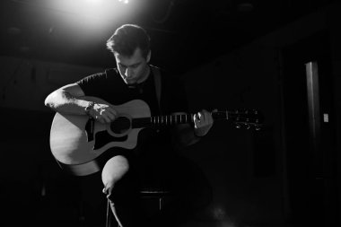 Avrupa görünümlü genç yakışıklı bir adam akustik gitar çalar ve bir sandalyeye oturur. Siyah beyaz fotoğraf..