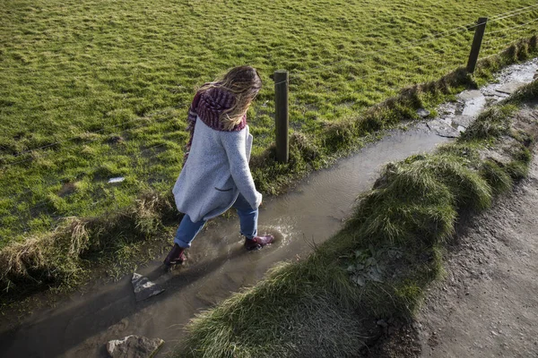 young woman with scarf coat and water boots walks through the puddles a cold autumn day in a green valley of ireland