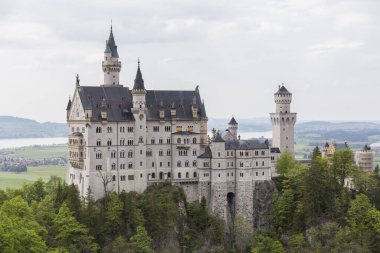 Neuschwanstein şatosunda fotoğraf çekimi, etkileyici Alman şatosuyla manzara, arka planda şatoyla poz veren insanlar, yeşil çayır, bol bitki örtüsü.