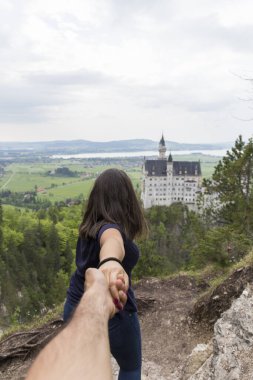 Neuschwanstein şatosunda fotoğraf çekimi, etkileyici Alman şatosuyla manzara, arka planda şatoyla poz veren insanlar, yeşil çayır, bol bitki örtüsü.