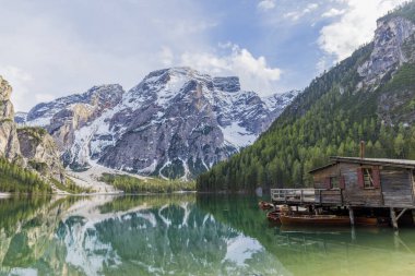 Iago di Braies manzarası, yüksek karlı dağları yansıtan etkileyici yeşil göl, bol yeşil bitki örtüsüyle çevrili, aynı zamanda iskele ve ahşap kayıklar olan ahşap bir kulübe görebiliriz.