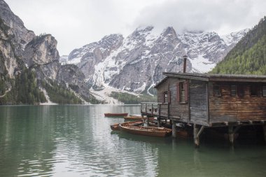 Iago di Braies manzarası, yüksek karlı dağları yansıtan etkileyici yeşil göl, bol yeşil bitki örtüsüyle çevrili, aynı zamanda iskele ve ahşap kayıklar olan ahşap bir kulübe görebiliriz.