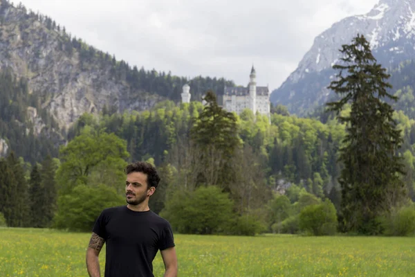 Neuschwanstein şatosunda fotoğraf çekimi, etkileyici Alman şatosuyla manzara, arka planda şatoyla poz veren insanlar, yeşil çayır, bol bitki örtüsü.