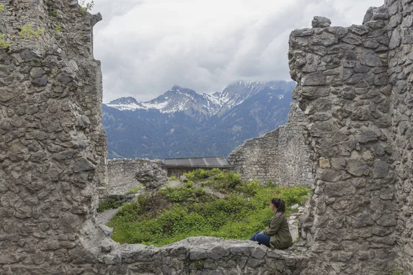 brunette woman with green jacket, sitting on stone ruins of ancient fortress watching the snowy mountains
