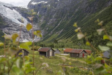 Terk edilmiş köy, farklı renkte ahşap evler, dağların arasında, arka planda buzullar, Norveç 'te bir yaz gününde bol miktarda yeşil bitki örtüsü.