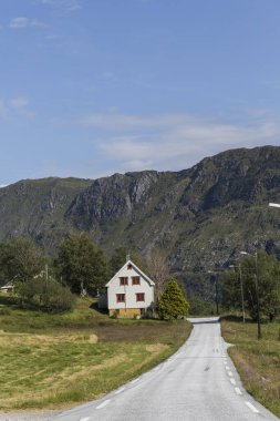 Sarı çiçeklerle dolu bir çayırdan geçen ve beyaz bir eve giden yol, arka planda dağları göreceksin, mavi gökyüzü ile aydınlık bir gün