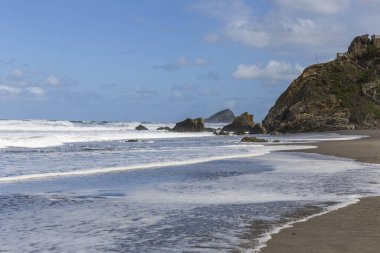 Koyu kumlu bir sahil manzarası, dalgalı deniz mavi gökyüzü, denizde kayalar.
