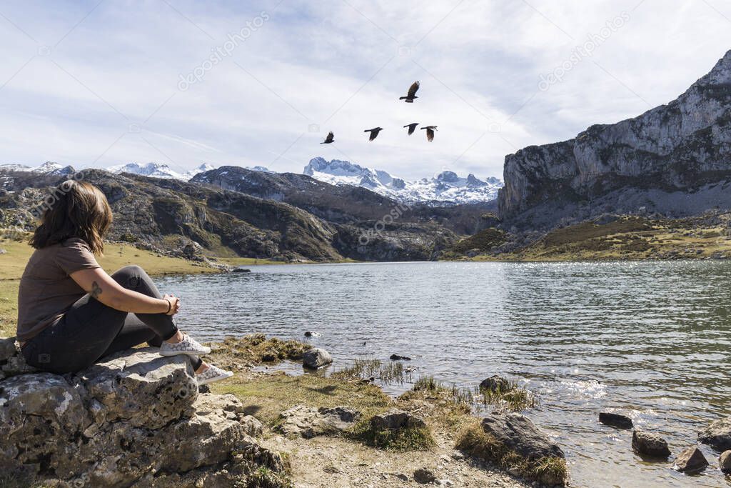 woman sitting on a rock on the shore of a lake between mountains looking at a flock of birds crossing in front
