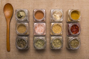 Değişik renkte baharatlar ve aromatik otlar ve çizgili cam kavanozlarda dokular, kahverengi arka planda, ahşap bir kaşıkla