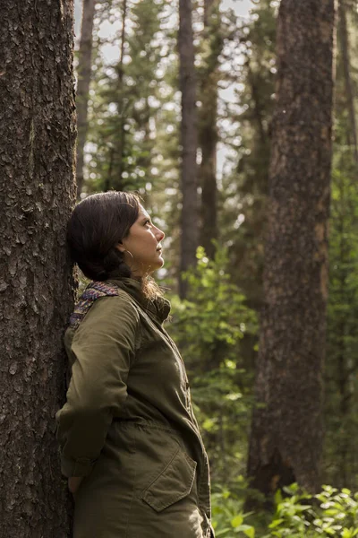 brunette woman with a braid and green jacket leans on the trunk of a forest tree