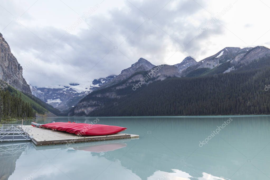 red canoes on a wooden jetty next to a turquoise water lake with high mountains next to it on a cloudy day