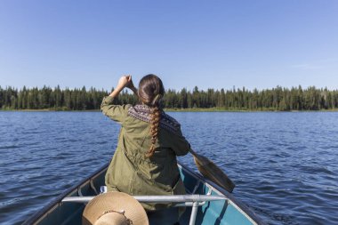 Tüylü ve yeşil ceketli esmer kadın, mavi su gölündeki ahşap bir teknede kürek çekiyor.