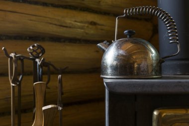 Demir bir şöminenin üzerinde metal çaydanlık ısınıyor, hemen yanında şöminenin aletlerini görebilirsiniz, duvar ahşap.