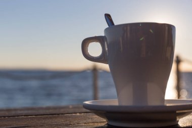 Ahşap bir masanın üzerindeki beyaz seramik bardağa yakın plan arkaplanda bulanık denizi görebilirsiniz.