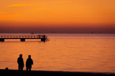 İsveç 'in Malmö şehrinde sıcak bir yaz akşamında gün batımına doğru yürüyen insanlar renkli deniz ve gökyüzüne doğru siluetler olarak görülür.