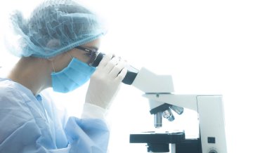 Beyaz arka planda mikroskopla virüsü analiz eden biyo-güvenlik kıyafetli İspanyol kadın doktor.