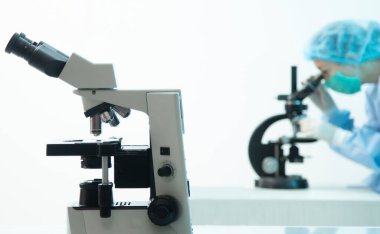 Ön planda optik mikroskop ve arka planda virüs analizi yapan kadın doktor.