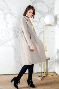 Parlak beyaz bir çatı katı tarzı stüdyosunda açık bej ceketli, kıvırcık saçlı güzellik mankeni kız. Güzel Lüks Kış Kadını. Güzellik ve moda. Bahar ve sonbahar kıyafetleri Stüdyoda poz veren kız
